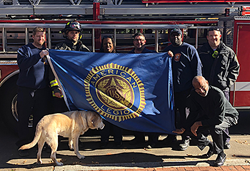 DC firemen and members of the American Legion stand with Galahad in front of a red firetruck and show off the Legion's new blue banner. Ever the food-oriented pup, he is focused on the treats in his dog walker Julie's pocket.