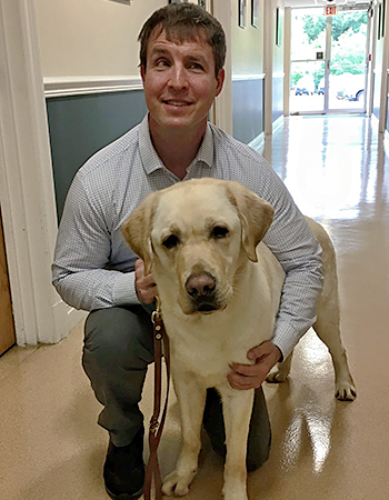 Having recently been matched as a team, Pat and Hogan smile for the camera in a hallway at the Guiding Eyes headquarters. Pat is kneeling next to standing Hogan, with his right arm around Hogan and right hand resting on Hogan's front leg.