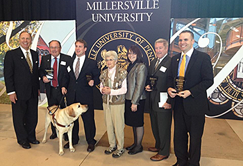 Appearing on stage with other recipients and University representatives, Pat receives the Young Alumni Achievement Award in the fall of 2014 at Millersville University. Galahad stands in front of the group and looks around.