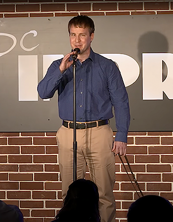 In the spring of 2018, Pat performs stand-up at DC Improv's graduation show. Outlined by the stage lights, the audience looks on as Pat stands with the mic in front of a red brick background and DC Improv's black sign.