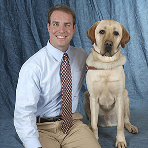 Pat, wearing a red and white checkered tie, sits with Galahad at the Guiding Eyes' graduation in front of an ocean blue backdrop.