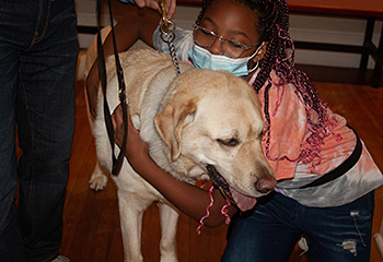 Inside the Workshop, a young girl gives Hogan a big hug. The happy pup stands, loving the attention, with his pink tongue partially hanging out of his mouth. The brown wood floor is seen below the pair.