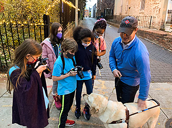 Five students direct their attention to Hogan. Two students point cameras at the pup while Pat, in a baseball cap and sky-blue half-zip pullover, holds Hogan's leash and gives him a pat on the back.