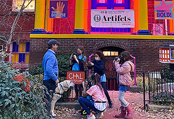 As evening sets in, five students gather around a working Hogan in front of the Capitol Hill Arts Workshop. The CHAW house is painted bright yellow and royal blue and illuminated with pink lights. Florescent orange letters C, H, A, and W adorn the building's four columns. Pat wears jeans, a blue sports pullover, and a Nationals baseball cap.