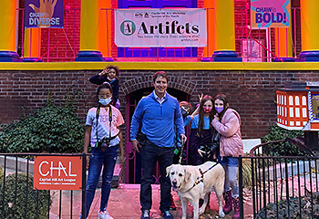 The happy gang poses for a group shot in front of the Capitol Hill Workshop. Hogan looks directly at the camera as the five students and Pat soak up these fun times together. One of the students playfully leans out from behind Pat and gives bunny ears to another student. The festive and brightly lit CHAW signage fills the background.