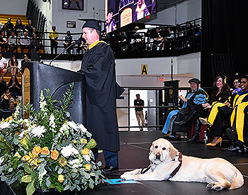 The shot is from stage left with Pat at the podium and guide dog Hogan attentively looking out at the crowd. Hogan is a yellow lab with an athletic build. Several people, wearing black gowns and caps and smiling, are seated behind Pat on stage. A portion of the crowd can be seen to the far left.