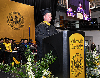 Taken from stage right, Pat delivers his speech. The stage party appears in the background to Pat’s left. A large yellow and white flower arrangement is on both sides of the podium. Pat’s image appears on a video screen at upper right of the photo.