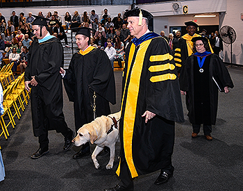 Moving at a quick clip, Pat and Josh enter the gymnasium with the stage party. Pat uses sighted guide with Josh while Hogan heals by their side. There are gray carpet and yellow chairs. The stage party wears black gowns and caps.