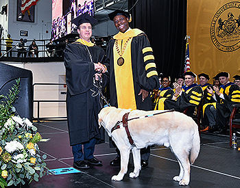 Pat shakes President Wubah’s hand following the remarks. The seated stage party is clapping behind them. Guide Hogan stands in front of the pair, looking at Pat. The black and yellow MU seal can be seen behind the stage party.