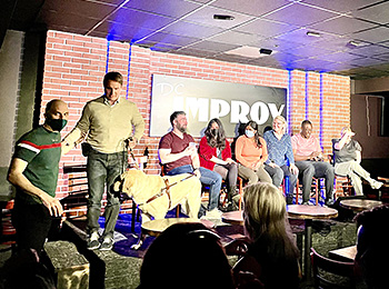 Pat and his classmates have just completed their graduation show. The team of seven is shown on the DC Improv main stage. Pat and Hogan are heading off the stage, with a helping hand from the instructor. Pat wears a tan V-neck sweater, dark jeans, and collared shirt. He instructs Hogan to heel as they prepare to head down the steps.
