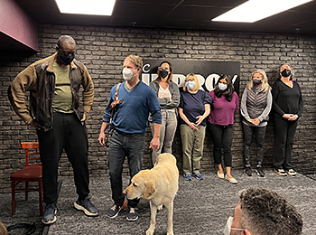 In the DC Improv lounge, the BAT class practices a Harold scene. Pat and his classmate have stepped to the forefront, downstage right. They are engaged in dialogue while yellow lab Hogan stands by Pat's side. The five remaining classmates wait their turn in the background. They're standing in front of a black brick wall that bears the DC Improv Comedy sign. Pat wears a blue V-neck sweater and jeans.