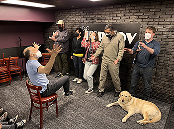 In the Improv lounge, Pat and five classmates are performing an animated skit on stage. Chris, their instructor, faces the students. He is perched precariously on a chair with both hands and one foot raised in the air. He's gesticulating wildly and the students, standing in front of a black brick wall with the DC Improv sign, motion enthusiastically back to him. Hogan, unphased, lies calmly at Pat's feet.