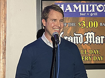 A bright spotlight centers on Pat as he performs a stand-up set. Pat, wearing a navy-blue sweater over a button-up shirt, is smiling into the mic. The sign for Hamilton's Bar and Grill can be seen in the background. Although not captured in the frame, working Hogan is taking a nap on Pat's foot.