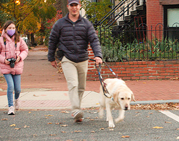 Hogan, a large yellow lab, guides Pat across the street on their way to the park. The guide dog team steps off the curb as a young photography student in a pink puffer jacket cradles her camera in the background. Pat sports a gray puffer jacket, khakis, and baseball cap.