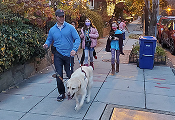 Four CHAW students are walking briskly in pursuit of a working Hogan. The kids are quickly moving with cameras around their necks as they attempt to catch Pat and Hogan. Pat is smiling and Hogan looks focused on his job. It's dusk while the happy bunch glides down the sidewalk together.