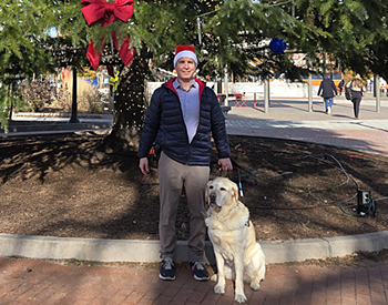 Pat and his yellow lab, Hogan, pause at the Eastern Market subway station on Capitol Hill in Washington, DC. The team stands in front of a festive green cedar tree adorned with red bows and snowflake lights. Pat wears a classic red-and-white Santa hat and a blue and red winter jacket.  Hogan, in harness, sits patiently at Pat's side. The Subway stop can be seen in the distance. Together, they take a brief, cheerful stop before heading off to start the workday.